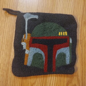 May include: A black felt square with a hand-stitched image of a Star Wars Boba Fett helmet. The helmet is green, red, and gray with yellow details.