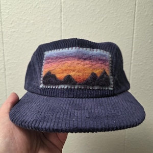 Peut inclure: Casquette en velours côtelé bleu marine avec un patch rectangulaire représentant un paysage de montagnes et un coucher de soleil aux tons orange, violet et bleu. La casquette a une visière incurvée.