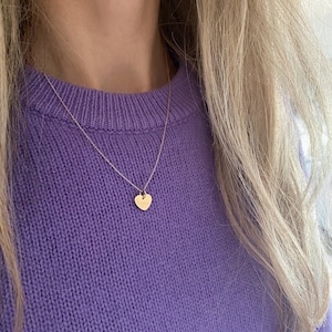 May include: A delicate gold chain necklace with a small heart pendant. The necklace is worn by a person with long blonde hair and a purple sweater.