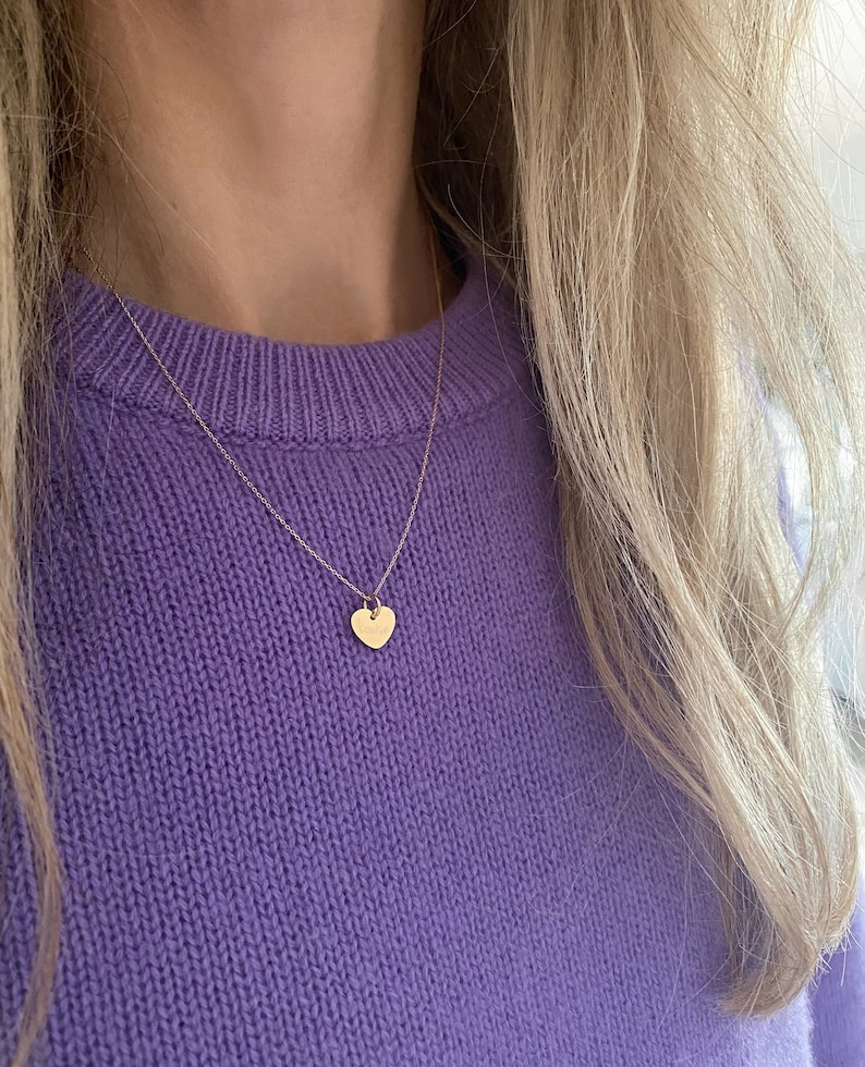 May include: A delicate gold chain necklace with a small heart pendant. The necklace is worn by a person with long blonde hair and a purple sweater.