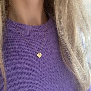 May include: A delicate gold chain necklace with a small heart pendant. The necklace is worn by a person wearing a purple sweater.