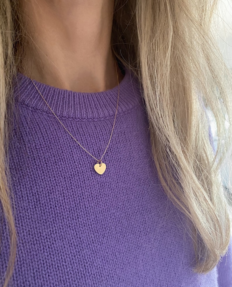 May include: A delicate gold chain necklace with a small heart pendant. The necklace is worn by a person wearing a purple sweater.