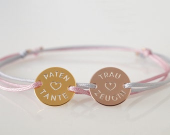 May include: Two friendship bracelets with gold and rose gold round charms. The gold charm says "PATEN TANTE" with a heart symbol. The rose gold charm says "TRAU ZEUGIN" with a heart symbol.