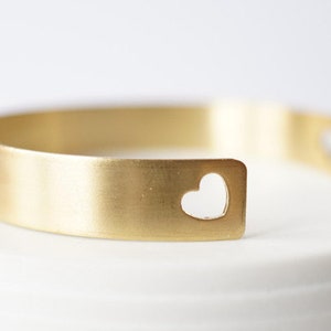 May include: A gold-coloured cuff bracelet with a heart-shaped cutout. The bracelet has a simple, minimalist design and a brushed metal finish. The heart cutout is centered on the front of the bracelet.
