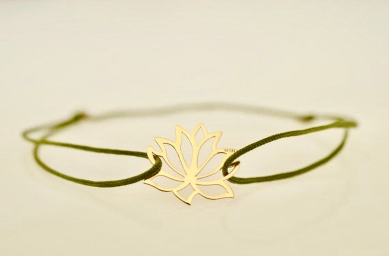 May include: A gold-colored lotus flower charm bracelet. The charm is a cutout design of the flower. The bracelet has a dark green cord. The bracelet is on a white background.