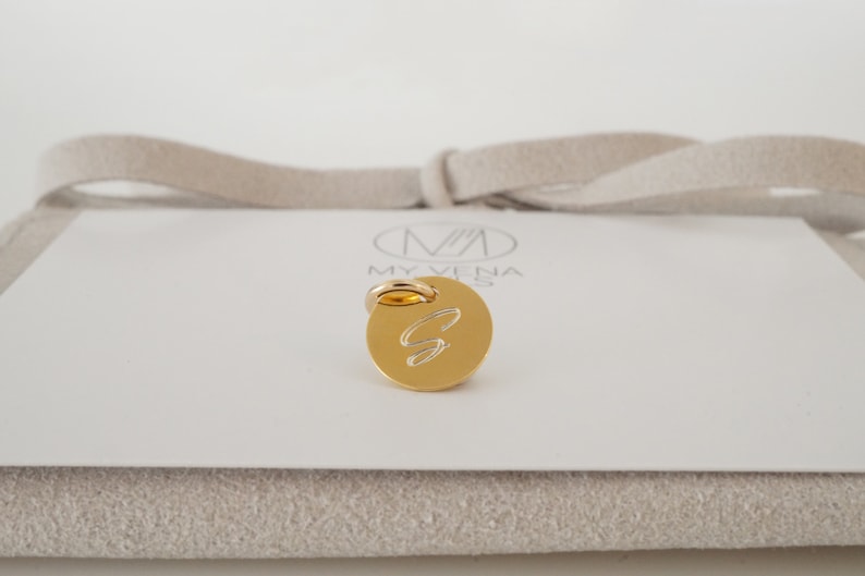May include: Gold charm with a cursive letter S engraved on a round disc. The charm has a ring attached to the top for attaching to a necklace or bracelet.