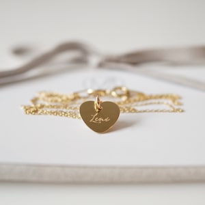 May include: A gold heart-shaped pendant necklace with the name "Lena" engraved on it. The necklace is on a white background with a beige suede box.