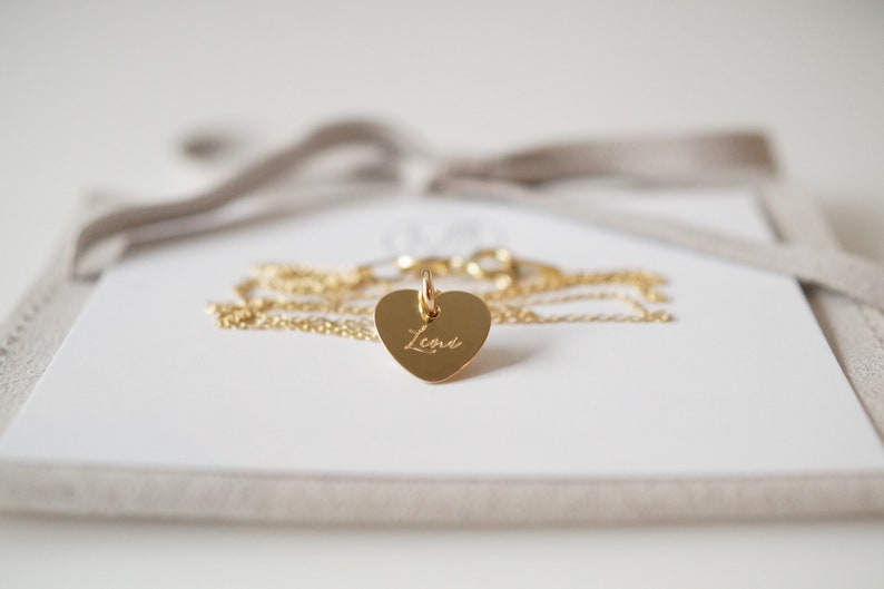 May include: A gold heart-shaped pendant necklace with the name "Lena" engraved on it. The necklace is on a white background with a beige suede box.