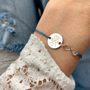 Könnte beinhalten: Ein blaues Kordelarmband mit einem silbernen Anhänger mit den Buchstaben B, C und A sowie einem Herzsymbol. Der Anhänger ist an einem Unendlichkeitszeichen befestigt.