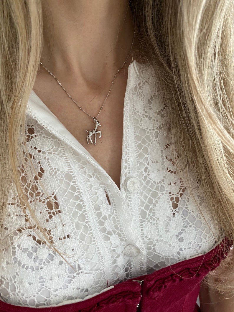 May include: A silver necklace with a deer pendant. The necklace is worn over a white lace blouse and a burgundy top. The deer pendant is a detailed representation of the animal.