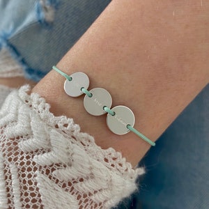 Könnte beinhalten: Ein hellblaues Armband aus Schnur mit drei runden Silberanhängern. Die Anhänger sind mit den Namen "Eva", "Anna" und "Fabian" graviert.