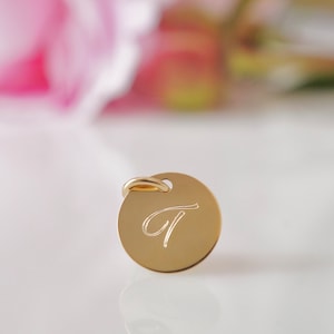 May include: A gold-colored round charm with a cursive letter "T" engraved on it. The charm has a small loop at the top for attaching to a necklace or bracelet.