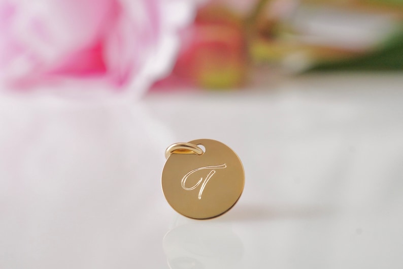May include: A gold-colored round charm with a cursive letter "T" engraved on it. The charm has a small loop at the top for attaching to a necklace or bracelet.
