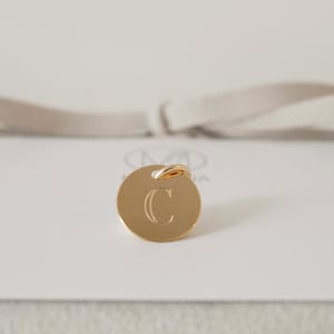 May include: A gold charm with a round disc featuring the letter "C" engraved in a serif font. The charm has a small gold loop at the top.