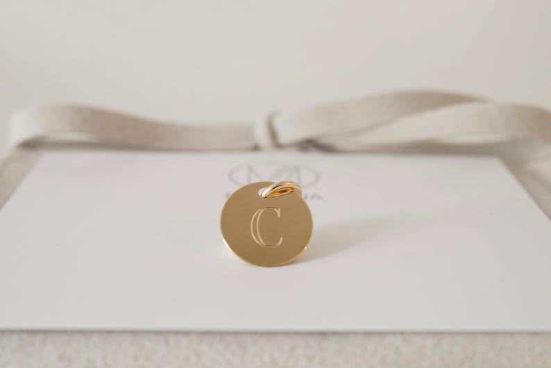 May include: A gold charm with a round disc featuring the letter "C" engraved in a serif font. The charm has a small gold loop at the top.