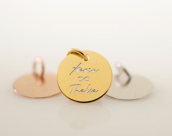 May include: A gold-colored round pendant with the names "Aaron" and "Thalia" engraved, separated by an infinity symbol. The pendant has a small loop at the top for a chain. Two other round pendants, one rose gold and one silver, are in the background.