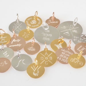 Op de afbeelding: Assortiment van ronde metalen bedels met verschillende gravures. Sommige bedels zijn goud, zilver en ros&eacute;goud. De bedels hebben tekst zoals "Aloha", "Leo Tatjana Anna", "fairy wander" en "Matteo 46 cm 2630 g 4.6.2020".