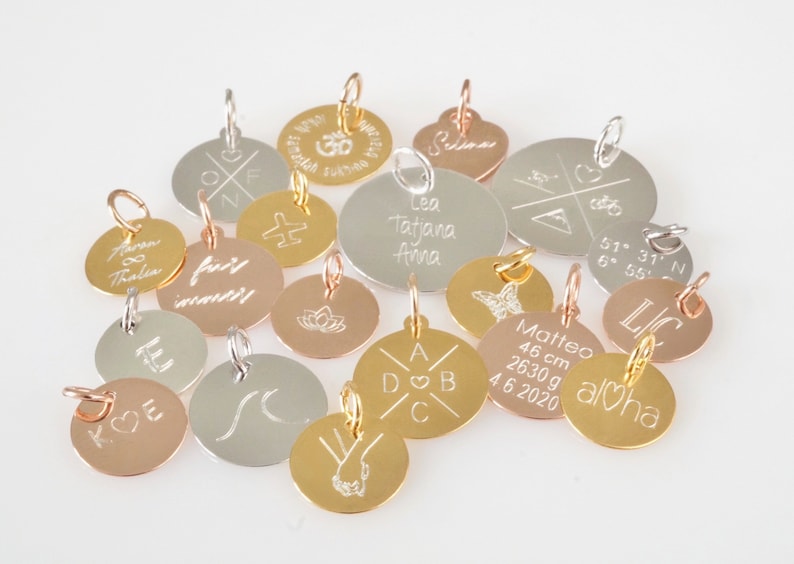Op de afbeelding: Assortiment van ronde metalen bedels met verschillende gravures. Sommige bedels zijn goud, zilver en ros&eacute;goud. De bedels hebben tekst zoals "Aloha", "Leo Tatjana Anna", "fairy wander" en "Matteo 46 cm 2630 g 4.6.2020".