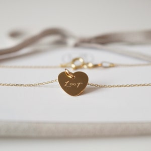 May include: A gold heart-shaped necklace with a delicate chain and a personalized inscription that reads "L&infin;T".
