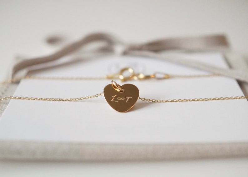 May include: A gold heart-shaped necklace with a delicate chain and a personalized inscription that reads "L&infin;T".