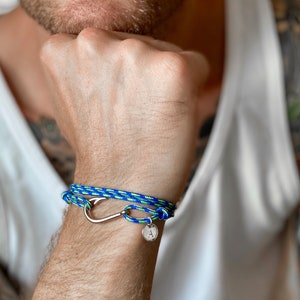 Könnte beinhalten: Ein geflochtenes Armband aus blauem und grünem Seil mit einem silbernen Haken-Anhänger und einem silbernen Scheiben-Anhänger mit dem Buchstaben 'A' darauf.