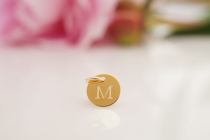 May include: A gold-colored charm with a round shape and a small loop at the top. The charm has the letter "M" engraved on the front.