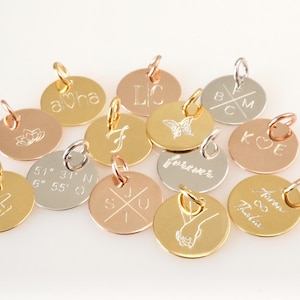 Op de afbeelding: Een verzameling van 12 ronde bedels van goud, zilver en ros&eacute;goud met verschillende gegraveerde ontwerpen, waaronder initialen, co&ouml;rdinaten, een vlinder, een hart, een kruis, een bloem en het woord "forever".