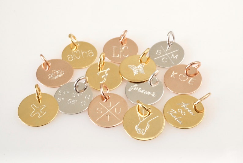 Op de afbeelding: Een verzameling van 12 ronde bedels van goud, zilver en ros&eacute;goud met verschillende gegraveerde ontwerpen, waaronder initialen, co&ouml;rdinaten, een vlinder, een hart, een kruis, een bloem en het woord "forever".