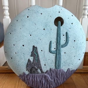 May include: A circular ceramic vase with a light blue speckled finish featuring a cutout design of a desert scene. The relief includes a dark gray coyote, a textured teal saguaro cactus, and a purple base. Small circular holes are scattered across the surface.