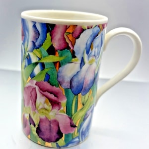 May include: A ceramic mug with a white handle and rim, featuring a floral design of irises in shades of purple, blue, and white. Green leaves and gold accents complete the design. The mug is cylindrical and stands upright.