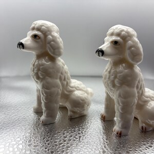 May include: Two white ceramic poodle figurines in a seated position. Each dog features a glossy finish, textured curly fur detailing, and painted black noses with brown eyes. The figures are identical in size, shape, and posture, facing left.