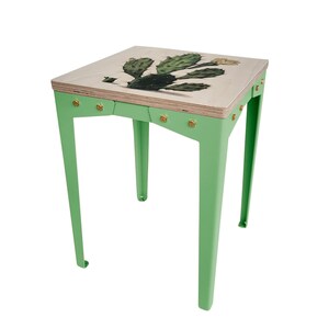 Op de afbeelding: Kleine, vierkante bijzettafel met een lichtgroen metalen frame en een houten blad. Het tafelblad heeft een gedetailleerde illustratie van een groene cactus met een gele bloem. De tafel is ongeveer 46 cm hoog.