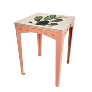Op de afbeelding: Een kleine vierkante bijzettafel met een licht perzikkleurig metalen frame en een houten blad. De bovenkant heeft een groene en witte cactusillustratie. De tafel is ongeveer 46 cm hoog en 41 cm breed.