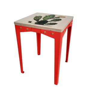 Op de afbeelding: Een kleine, vierkante bijzettafel met een rood metalen frame en een houten blad. Het tafelblad heeft een gedetailleerde illustratie van een groene cactus met een gele bloem. De tafel is ongeveer 46 cm hoog.