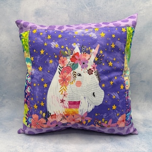 Könnte beinhalten: Quadratisches Zierkissen mit einem weißen Einhorn mit Blumenkrone auf einem lila Sternenhintergrund. Das Kissen hat einen lila gepunkteten Rand und farbenfrohe Seitenteile. Maße ca. 40 cm x 40 cm.