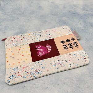 Könnte beinhalten: Eine weiße Reißverschlusstasche mit einem bunten Spritzmuster. Die Tasche ist in vier Abschnitte mit verschiedenen Mustern unterteilt, darunter ein rosa Waschbär, ein rosa-weißes Sternmuster und ein rosa Abschnitt mit schwarzen Blumen. Die Tasche hat ein Etikett mit der Aufschrift "Happy"