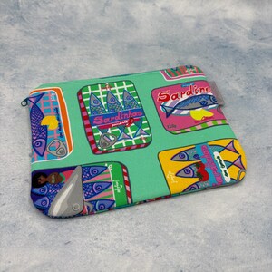 Könnte beinhalten: Eine rechteckige, mit einem Reißverschluss versehene Tasche mit türkisfarbenem Hintergrund. Die Tasche ist mit farbenfrohen Sardinenbüchsen-Illustrationen bedeckt. Die Dosen zeigen verschiedene Designs mit Fischen, Text und leuchtenden Farben wie Pink, Gelb und Blau.