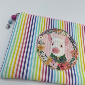 Könnte beinhalten: Eine regenbogenfarbene, gestreifte Kosmetiktasche mit Reißverschluss und einem rosa Kreis, in dem ein Schwein mit einem Blumenkranz und einem blau-grünen gemusterten Hemd zu sehen ist.