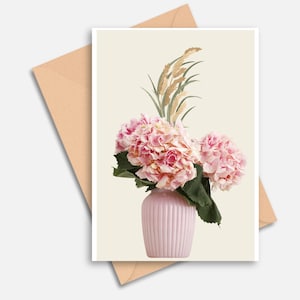 Pode incluir: Um cartão com um vaso rosa cheio de flores de hortênsia rosa e gramíneas ornamentais. O cartão está em um fundo creme, com um envelope marrom ao fundo. O vaso tem ranhuras verticais.