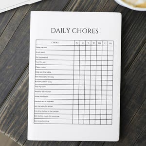 Pode incluir: Um bloco de notas branco com as palavras "DAILY CHORES" no topo. Abaixo, uma grade lista tarefas diárias como "Fazer a cama", "Escovar os dentes" e "Fazer o dever de casa". O bloco de notas está sobre uma superfície de madeira escura.
