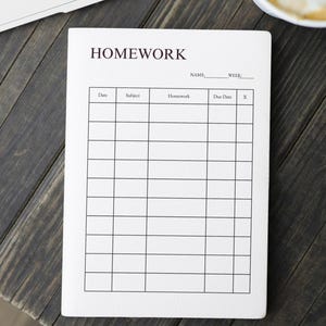 Op de afbeelding: Een wit huiswerkboek met het woord 'HOMEWORK' bovenaan. Het boek heeft een raster voor het invullen van data, vakken, huiswerkopdrachten, vervaldatums en een afvinkkolom. Het boek ligt op een donker houten oppervlak.