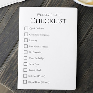 Pode incluir: Um bloco de notas branco com a frase "CHECKLIST DE REINÍCIO SEMANAL" impressa. A lista inclui tarefas como "Quick Declutter" e "Digital Detox (1 hora)". O bloco de notas está sobre uma superfície de madeira escura.