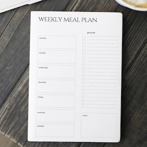 Pode incluir: Um bloco de notas branco para um plano de refeições semanal com o texto "WEEKLY MEAL PLAN" no topo. O bloco de notas tem secções para cada dia da semana, uma lista de compras e notas. O bloco de notas está numa superfície de madeira escura.