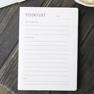 Pode incluir: Um bloco de notas branco com as palavras "TO DO LIST" no topo, e linhas para "TOP PRIORITIES" e "TO DO LIST" abaixo. O bloco de notas está sobre uma superfície de madeira escura.