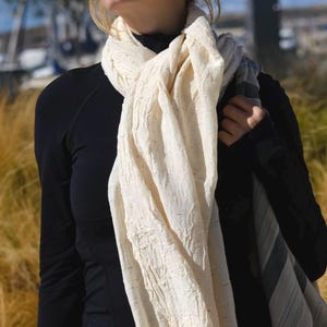 May include: A cream-colored scarf with fringe is draped over the shoulder. The scarf has a textured appearance and is paired with a black long-sleeve top and black leggings. The background features tall grass and a blue sky.