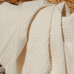 May include: A cream-coloured waffle weave throw blanket with fringe edges is folded and placed in a woven wicker basket.