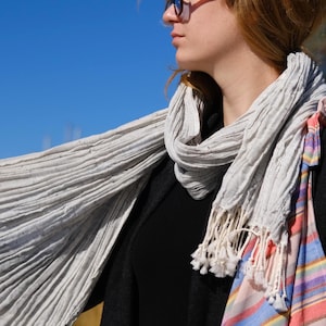 May include: A light gray, pleated scarf with white fringe is draped over a black top and coat. A striped tote bag with pink, blue, and orange hues hangs on the shoulder. The background is a clear blue sky.