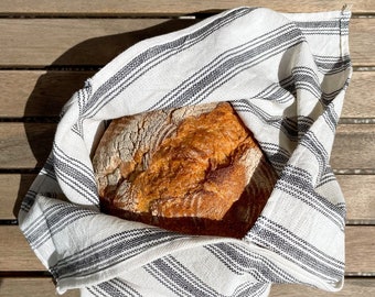 May include: A freshly baked loaf of bread wrapped in a white and black striped kitchen towel. The bread is golden brown and has a crusty exterior.