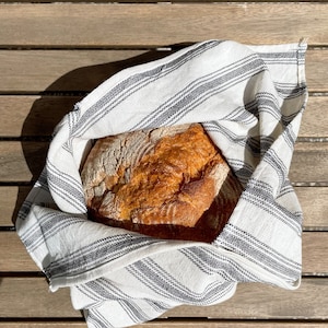 Op de afbeelding: Een vers gebakken brood gewikkeld in een wit-zwarte gestreepte theedoek. Het brood is goudbruin en heeft een knapperige korst.