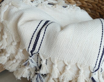 May include: A white throw blanket with navy blue stripes and fringe. The blanket is folded and placed in a woven basket.